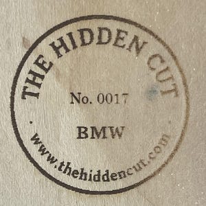 May include: Circular stamp with the text "THE HIDDEN CUT" and "No. 0017" in a dark brown color. The word "BMW" is printed below the number, and the website address "www.thehiddencut.com" is at the bottom.