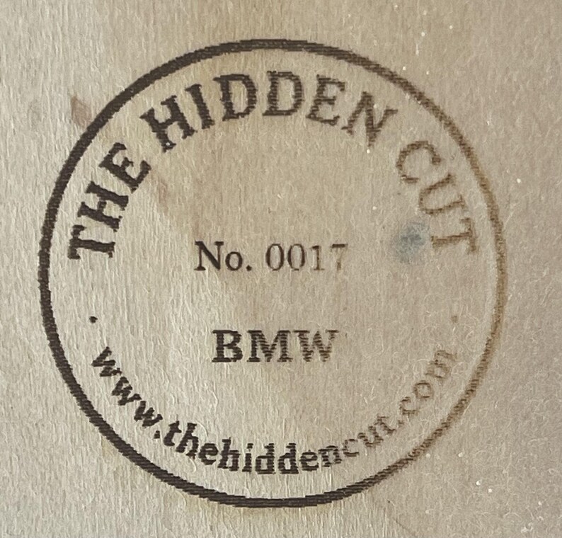 May include: Circular stamp with the text "THE HIDDEN CUT" and "No. 0017" in a dark brown color. The word "BMW" is printed below the number, and the website address "www.thehiddencut.com" is at the bottom.