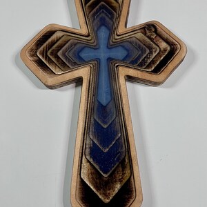 May include: Wooden cross with a layered design. The cross features a light brown outer layer, with a blue and brown gradient in the center. The cross is a religious symbol.