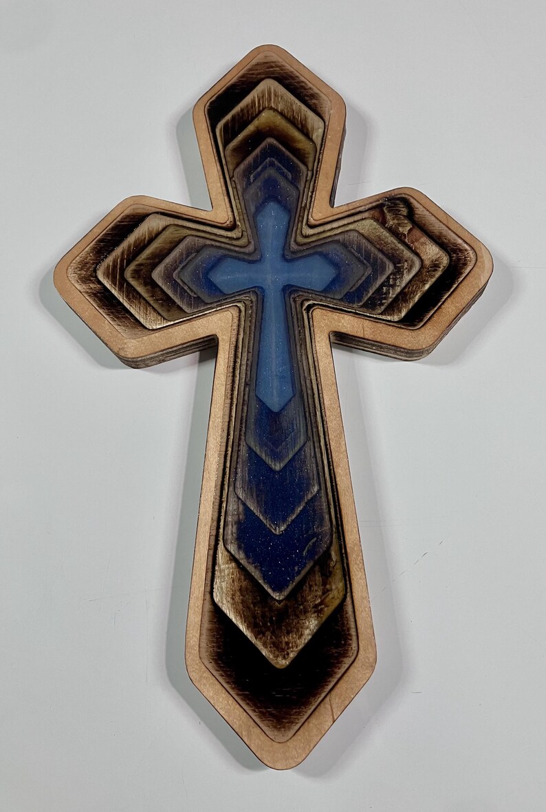 May include: Wooden cross with a layered design. The cross features a light brown outer layer, with a blue and brown gradient in the center. The cross is a religious symbol.