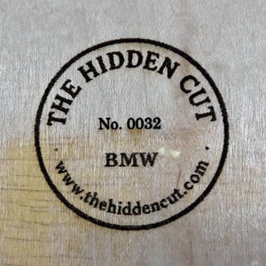 May include: Circular stamp on a light wood surface. The stamp reads "THE HIDDEN CUT" around the top, "No. 0032" in the center, and "BMW" below. The website address www.thehiddencut.com is at the bottom.