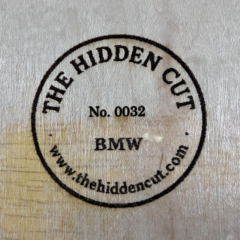 May include: Circular stamp on a light wood surface. The stamp reads "THE HIDDEN CUT" around the top, "No. 0032" in the center, and "BMW" below. The website address www.thehiddencut.com is at the bottom.