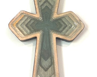 May include: A layered wooden cross with a geometric design. The cross features a light brown border, with shades of gray and green within the layers. The cross is approximately 4 inches tall.