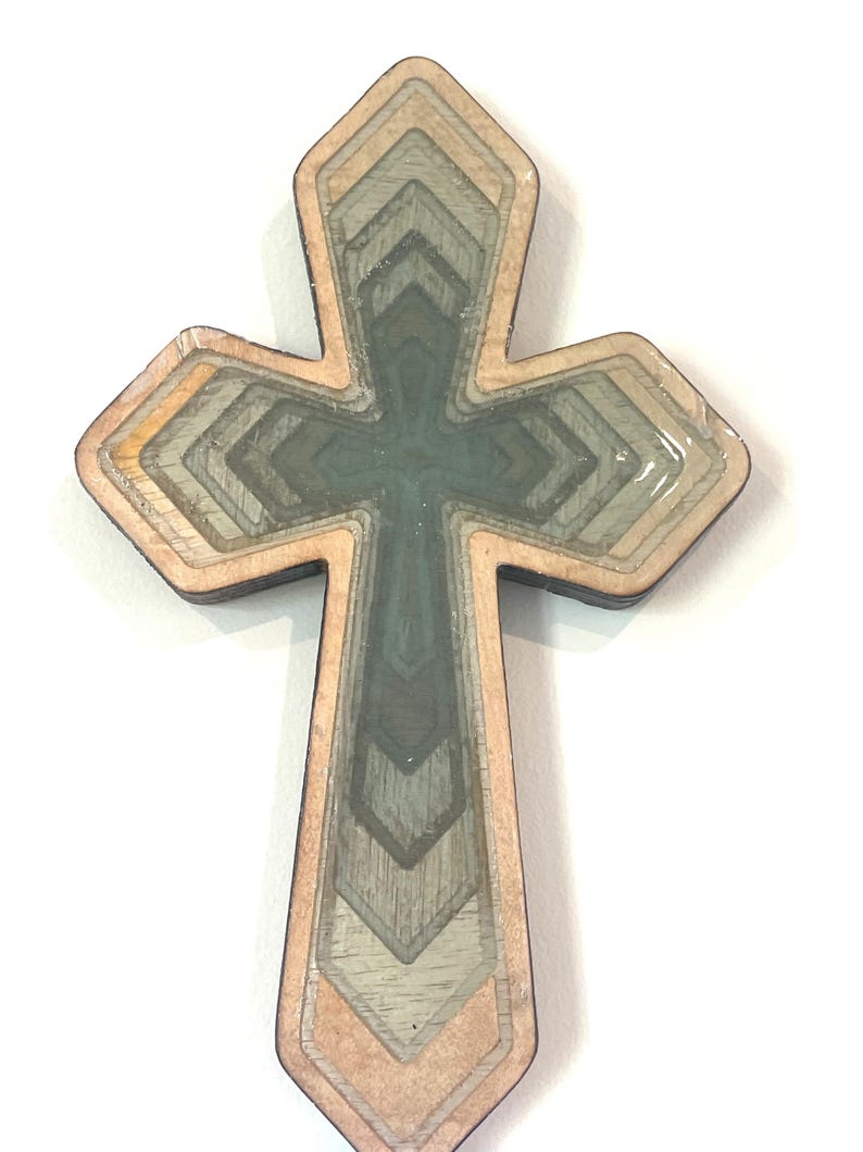 May include: A layered wooden cross with a geometric design. The cross features a light brown border, with shades of gray and green within the layers. The cross is approximately 4 inches tall.