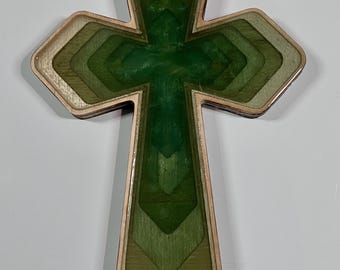 May include: Wooden cross with a layered design. The cross features a central section of deep green, surrounded by lighter shades of green and a natural wood border. The cross is a religious symbol.