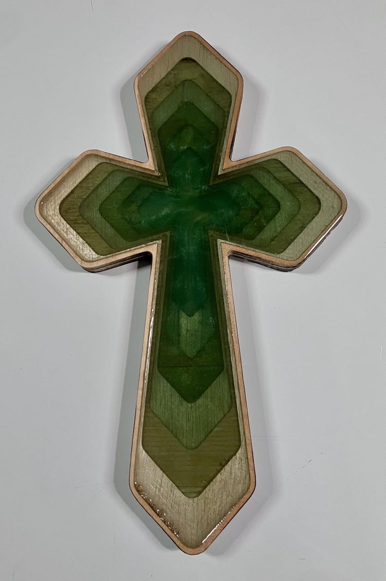 May include: Wooden cross with a layered design. The cross features a central section of deep green, surrounded by lighter shades of green and a natural wood border. The cross is a religious symbol.