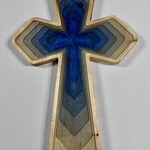 May include: Wooden cross with a layered design. The cross features a blue and silver resin center, surrounded by graduated wooden layers. The outer edges are a natural wood color.