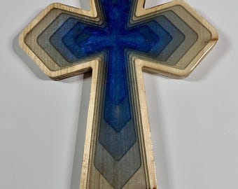 May include: Wooden cross with a layered design. The cross features a blue and silver resin center, surrounded by graduated wooden layers. The outer edges are a natural wood color.
