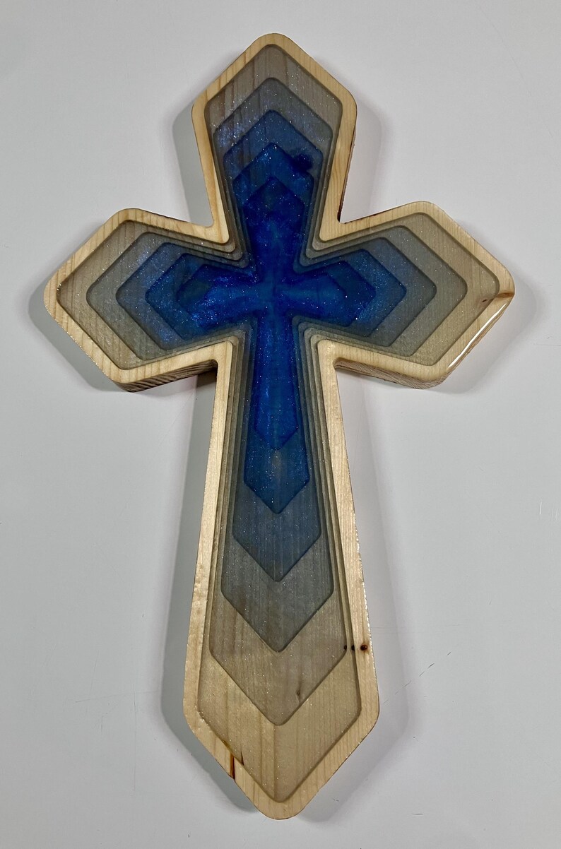 May include: Wooden cross with a layered design. The cross features a blue and silver resin center, surrounded by graduated wooden layers. The outer edges are a natural wood color.