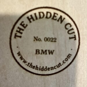 May include: A circular wooden emblem with the text "THE HIDDEN CUT" in a curved font. Below, it reads "No. 0022" and "BMW". The website address "www.thehiddencut.com" is also visible. The emblem is a light brown color.