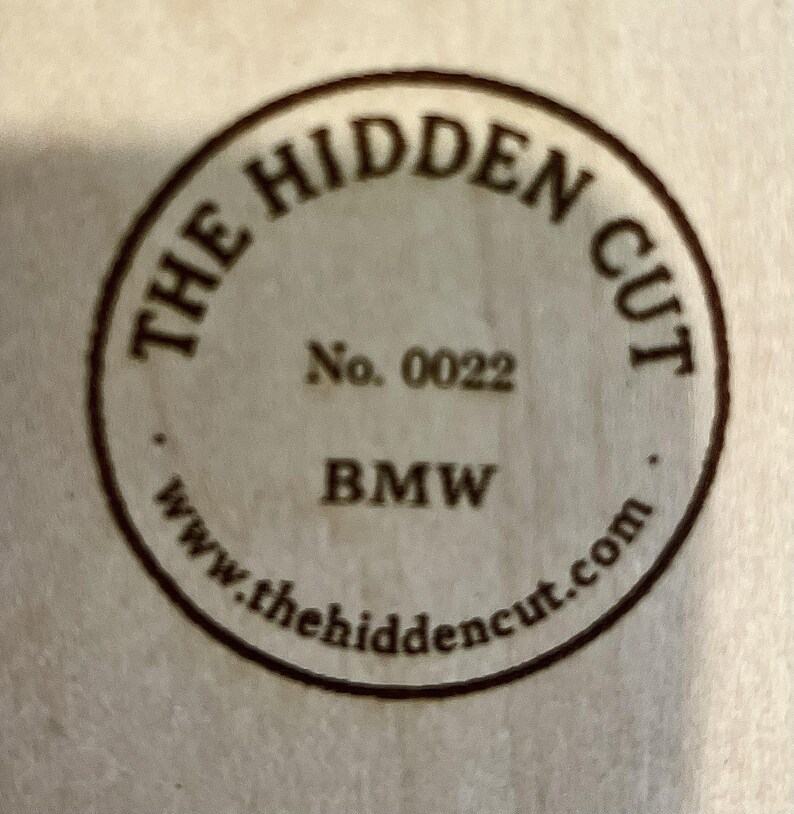 May include: A circular wooden emblem with the text "THE HIDDEN CUT" in a curved font. Below, it reads "No. 0022" and "BMW". The website address "www.thehiddencut.com" is also visible. The emblem is a light brown color.