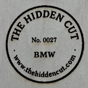 May include: A circular black stamp with the text "THE HIDDEN CUT" around the top and "www.thehiddencut.com" around the bottom. The stamp also includes "No. 0027" and "BMW" in the center.