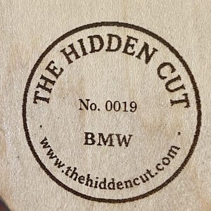 May include: A circular wooden stamp with the text "THE HIDDEN CUT" in a brown, circular border. The stamp also includes "No. 0019", "BMW", and the website "www.thehidencut.com".