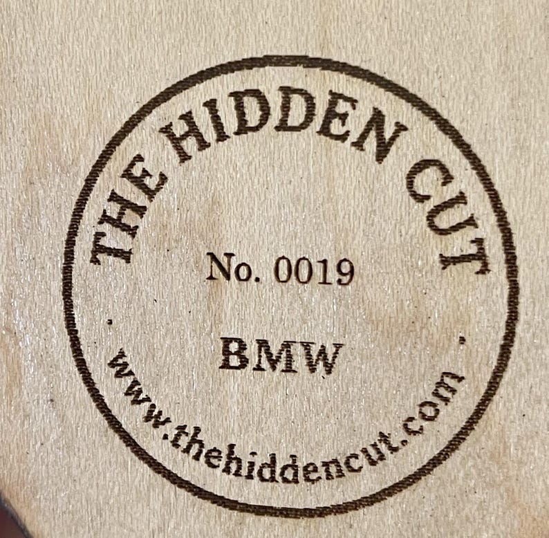 May include: A circular wooden stamp with the text "THE HIDDEN CUT" in a brown, circular border. The stamp also includes "No. 0019", "BMW", and the website "www.thehidencut.com".