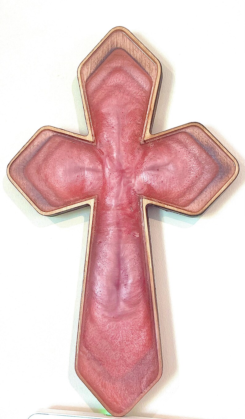 May include: A decorative cross with a wooden frame and a pink resin center. The cross has a layered design with a lighter pink hue in the center and a darker pink outline. The wooden frame adds a natural contrast.