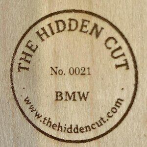 May include: Circular wooden stamp with the text "THE HIDDEN CUT" in a ring around the edge. The text "No. 0021" is centered, with "BMW" below it. The website "www.thehiddencut.com" is at the bottom.