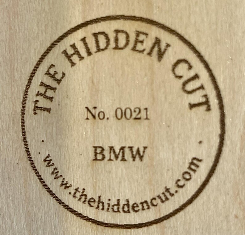 May include: Circular wooden stamp with the text "THE HIDDEN CUT" in a ring around the edge. The text "No. 0021" is centered, with "BMW" below it. The website "www.thehiddencut.com" is at the bottom.