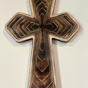 May include: A layered wooden cross with a detailed design. The cross features a series of nested shapes, creating a three-dimensional effect. The wood has a natural, warm tone with darker accents, and the edges are smooth.
