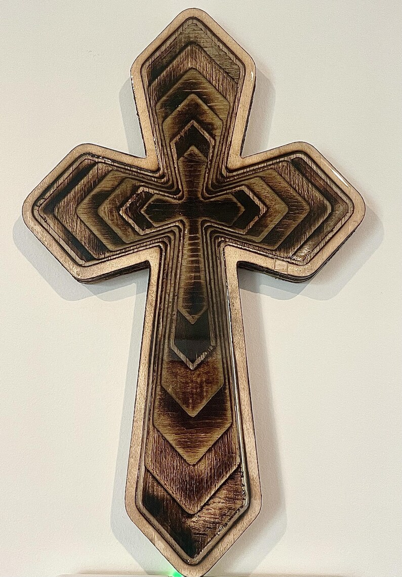 May include: A layered wooden cross with a detailed design. The cross features a series of nested shapes, creating a three-dimensional effect. The wood has a natural, warm tone with darker accents, and the edges are smooth.