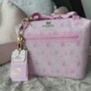 Peut inclure: Un sac &agrave; main rose clair avec un motif r&eacute;p&eacute;titif de petites ic&ocirc;nes blanches et le texte "Juicy Couture" imprim&eacute; sur le devant. Le sac comprend une attache en cha&icirc;ne m&eacute;tallique dor&eacute;e avec une &eacute;tiquette de bagage rectangulaire assortie.