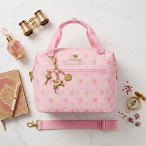 Peut inclure: Un sac &agrave; d&eacute;jeuner rose clair &agrave; motif floral, portant l'inscription Stanley LoveShackFancy, un ensemble de breloques dor&eacute;es avec un c&oelig;ur, un cheval ail&eacute; et un embl&egrave;me circulaire, une bandouli&egrave;re amovible, un flacon de parfum, des jumelles et un livre.