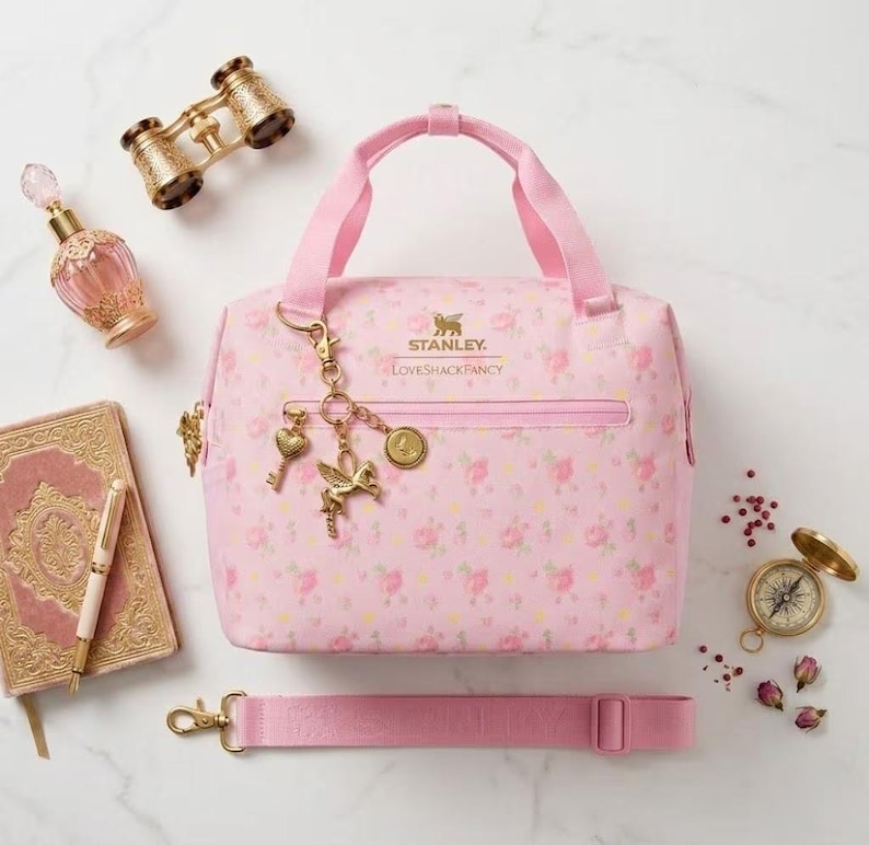 Peut inclure: Un sac &agrave; d&eacute;jeuner rose clair &agrave; motif floral, portant l'inscription Stanley LoveShackFancy, un ensemble de breloques dor&eacute;es avec un c&oelig;ur, un cheval ail&eacute; et un embl&egrave;me circulaire, une bandouli&egrave;re amovible, un flacon de parfum, des jumelles et un livre.