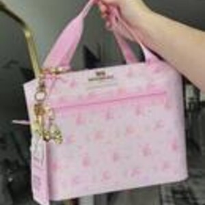Peut inclure: Un sac &agrave; main rose clair avec un motif r&eacute;p&eacute;titif de petites ic&ocirc;nes rose fonc&eacute;. Le sac poss&egrave;de deux anses, une poche frontale zipp&eacute;e horizontale et une breloque en m&eacute;tal dor&eacute; attach&eacute;e sur le c&ocirc;t&eacute;. Le texte "MINISO" est imprim&eacute; sur le devant.