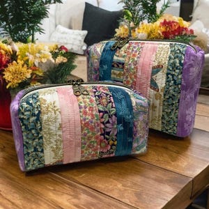 May include: Two quilted cosmetic bags with floral and patterned fabric panels in shades of pink, blue, green, and purple. The bags have a zipper closure and are displayed on a wooden table. The background includes flowers and a sofa.