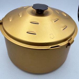 May include: A round, metallic gold cooking pot with a matching lid. The lid features a dark brown, scalloped-edge knob at the center, several small curved ventilation slits arranged in a circular pattern, and a small arrow symbol pointing right.