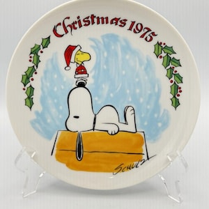 May include: A decorative plate featuring Snoopy lying on his doghouse, with Woodstock wearing a Santa hat. The plate has the text "Christmas 1975" and is decorated with holly and snow. Signed "Schulz".