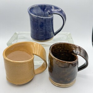 May include: Three ceramic mugs with a unique design where the handle is integrated into the rim. One mug is dark blue, one is a light tan, and one is a dark brown. Each mug has a smooth, glazed finish and a cylindrical body with a flat base.