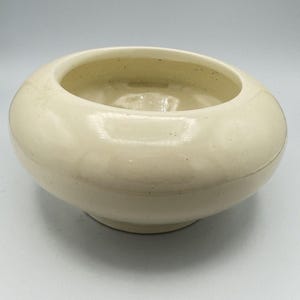 Puede incluir: Un cuenco de cerámica de color crema con forma redondeada y una amplia abertura. El cuenco se asienta sobre una pequeña base circular. El interior del cuenco es visible y la superficie tiene un acabado brillante. Este cuenco decorativo es adecuado para la decoración del hogar.