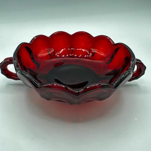 Könnte beinhalten: Eine rubinrote Glasschale mit gewelltem Rand und zwei kleinen Griffen. Die Schale hat eine tiefe, satte Farbe und eine glänzende Oberfläche. Das Design erinnert an Vintage-Glaswaren.