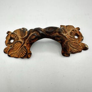 May include: Ornate, antique-style drawer pull with a curved handle. The handle is dark brown, and the decorative ends feature a leaf design in shades of orange and brown. The hardware is made of metal and has a distressed finish.