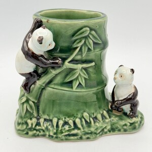 May include: A green ceramic cylindrical holder shaped like a bamboo stalk, featuring two small panda figures. One panda is climbing the side of the bamboo, while the other sits at the base. The piece has a glossy finish and detailed textures.