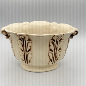 Puede incluir: Jardinera de cerámica color crema con borde festoneado y asas decorativas. La jardinera presenta paneles verticales con diseños de hojas marrones. La base tiene una forma ligeramente elevada y redondeada.