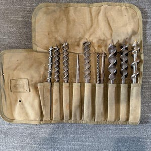 May include: A set of eight metal auger drill bits of varying diameters and designs, organized in a tan canvas tool roll with individual stitched pockets. The bits feature spiral flutes and pointed tips, arranged horizontally within the fabric slots.