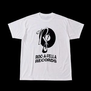 May include: A white short-sleeve t-shirt featuring a black graphic print on the chest. The design includes a stylized letter R integrated with a circular record shape and the text ROC-A-FELLA RECORDS printed in a bold, sans-serif font below it.