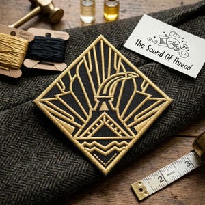 May include: A black and gold embroidered patch with an art deco design. The patch is square with a geometric pattern and the text "The Sound Of Thread" on a white card. Other items include spools of thread, a hammer, and a measuring tape.