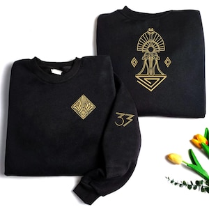 May include: Two black sweatshirts with gold geometric designs. One has a diamond-shaped design on the chest and the number "33" on the sleeve. The other features a larger, more elaborate design in gold.