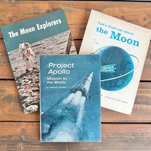 May include: Three vintage books on a wooden surface. The books are titled "The Moon Explorers", "Project Apollo", and "Let's find out about the Moon". The covers feature illustrations of astronauts, rockets, and the moon.