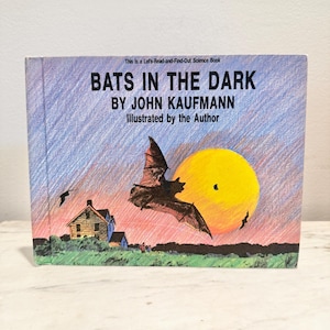 May include: A children's book titled "Bats in the Dark" by John Kaufmann. The cover features a bat in flight against a yellow sun and a sunset sky. The book is a science book for children.