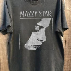 Pode incluir: T-shirt cinza escuro com o nome da banda "MAZZY STAR" acima de uma imagem em tons de cinza de um perfil humano dentro de um retângulo branco. A camiseta está pendurada em um cabide de madeira.