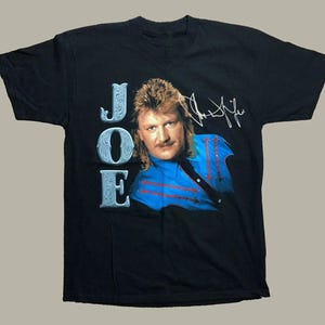 May include: Black t-shirt featuring a portrait of a man with long hair and a mustache, wearing a blue shirt with red accents. The word "JOE" is printed vertically on the left side of the shirt in a decorative font, and a signature is visible on the upper right.