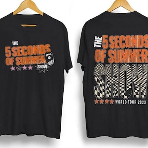 Puede incluir: Camiseta negra con el texto "THE 5 SECONDS OF SUMMER SHOW" en naranja y blanco. La parte delantera presenta un dibujo animado y estrellas. La parte trasera de la camiseta muestra "WORLD TOUR 2023".