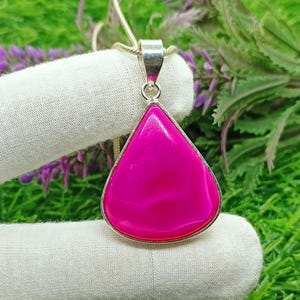 Puede incluir: Colgante en forma de lágrima con una piedra rosa vibrante engastada en un marco de metal plateado. La piedra pulida presenta sutiles variaciones de color. El colgante cuelga de una delicada cadena plateada, una joya llamativa.