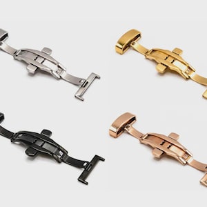 May include: Four watch clasps in different metallic finishes: silver, gold, black, and rose gold. Each clasp is a butterfly style, designed to secure watch bands. The clasps are displayed against a light background.