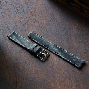 Könnte beinhalten: Ein grau-meliertes Leder-Uhrenarmband mit weißen Nähten und einer silberfarbenen Schnalle. Das Armband ist in zwei Teilen auf einer dunklen Holzoberfläche dargestellt. Das Leder hat ein abgenutztes, strukturiertes Aussehen.