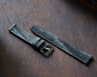 May include: A distressed gray leather watch strap with white stitching and a silver-colored buckle. The strap is shown in two pieces, lying on a dark wood surface. The leather has a worn, textured appearance.