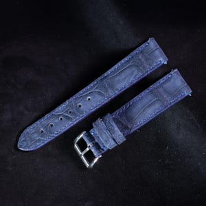 Puede incluir: Correa de reloj de dos piezas en un tono azul oscuro, con un patrón de cocodrilo texturizado. La correa está cosida con hilo azul en contraste e incluye una hebilla plateada. La correa de cuero está diseñada para un reloj clásico.
